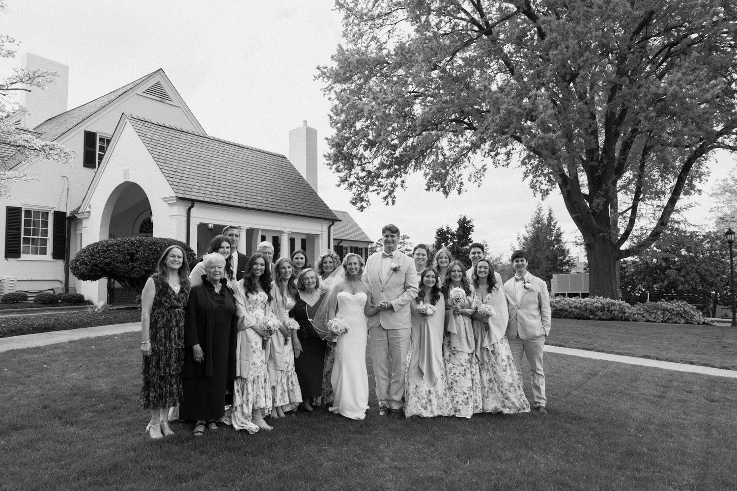 A black and white wedding party poses together on a lawn in front of a white church building with trees in the background.