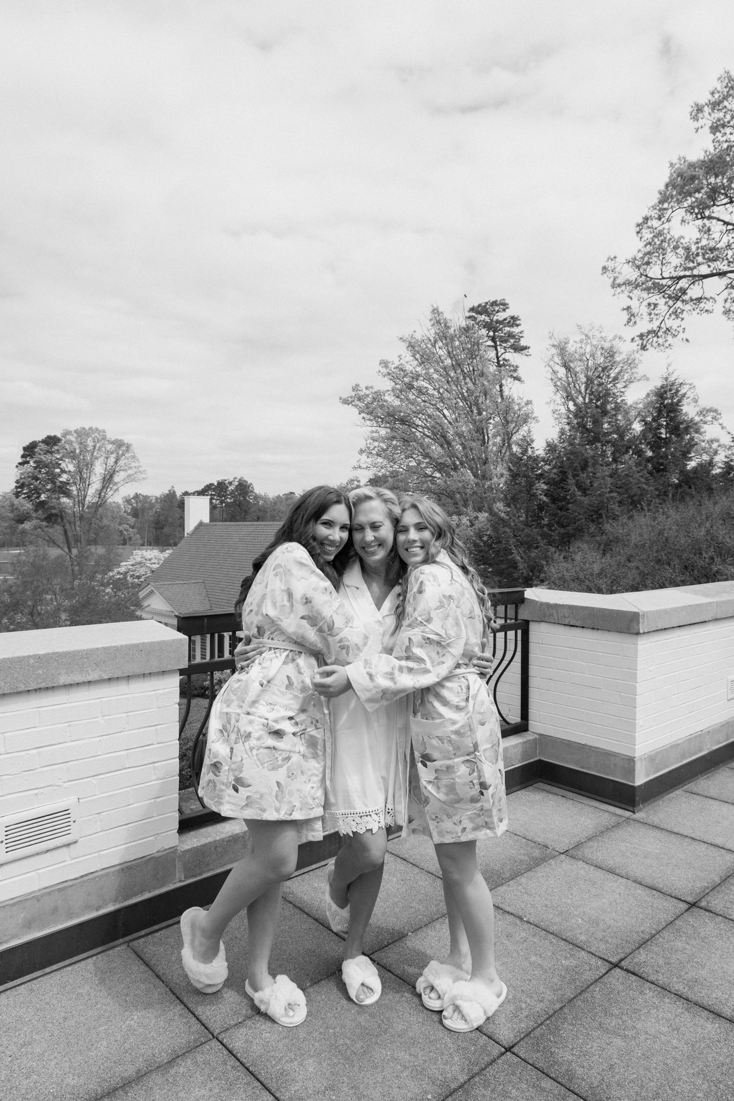 Three figures in matching floral robes and slippers share a warm embrace on a rooftop terrace on a cloudy day.