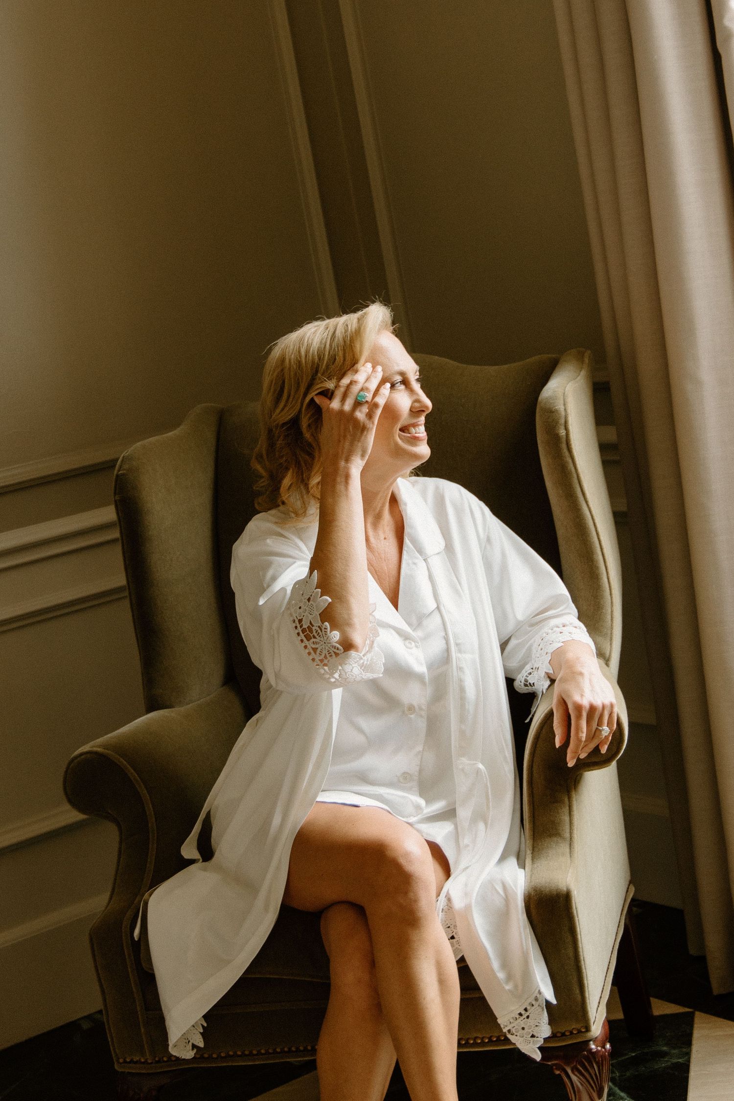 A person in a white robe sits elegantly in a vintage armchair by a curtained window in warm lighting.