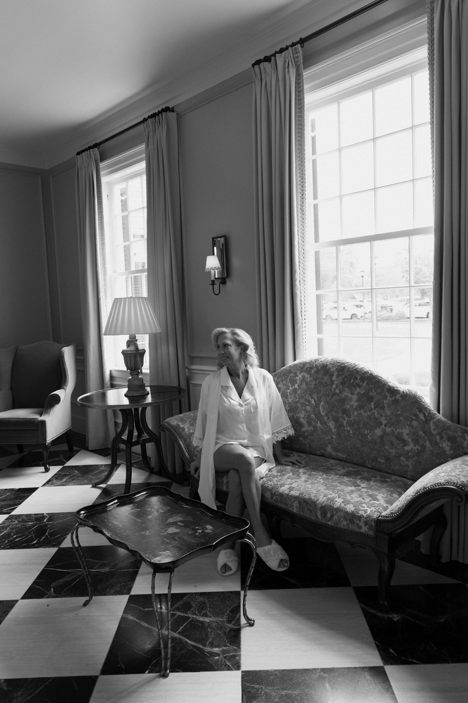 Black and white interior photo showing a living room with checkerboard floor, vintage furniture and tall windows with drapes.