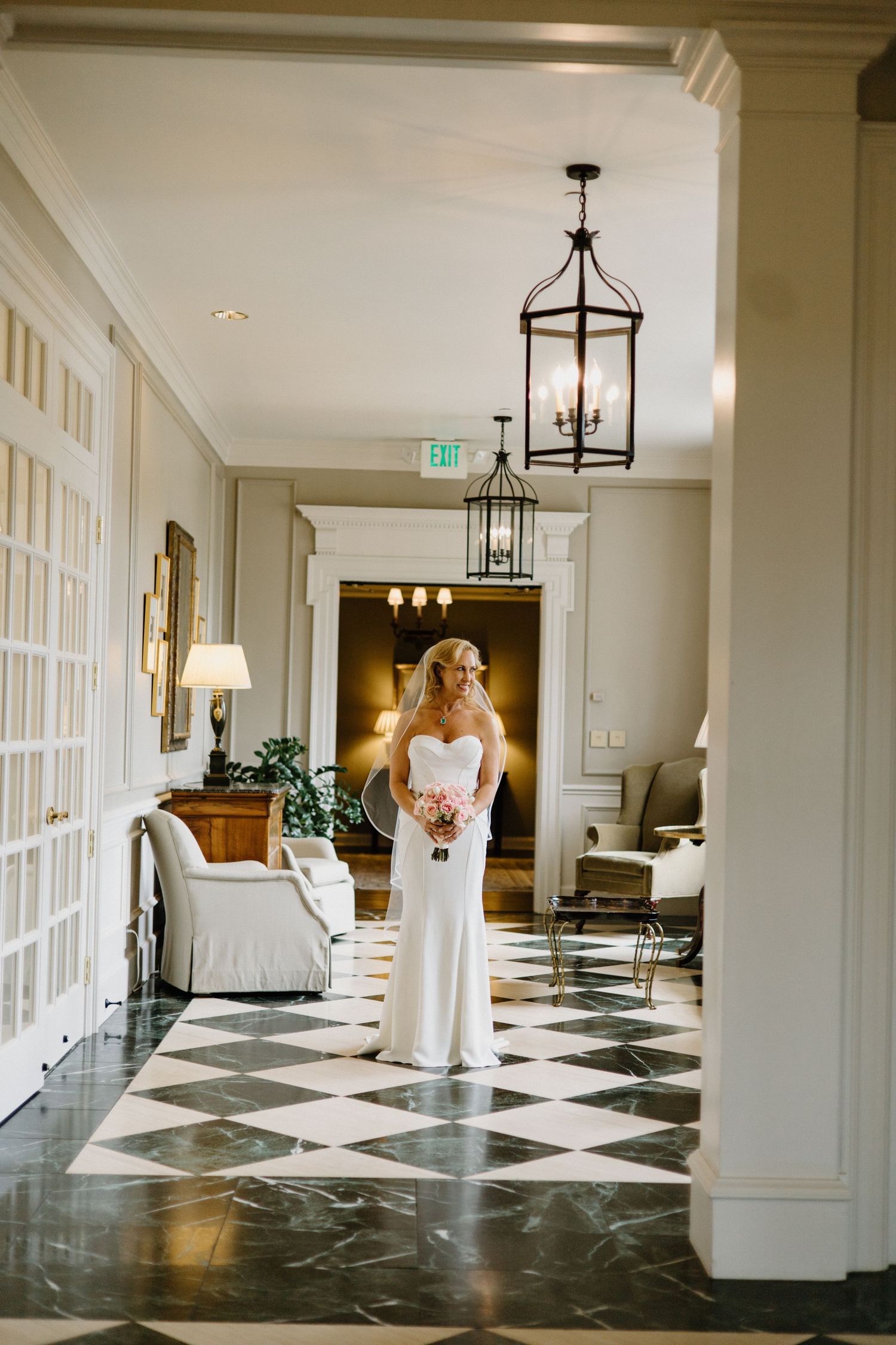 A figure in an elegant white wedding gown stands in an elegant hallway with black and white checkered floors.