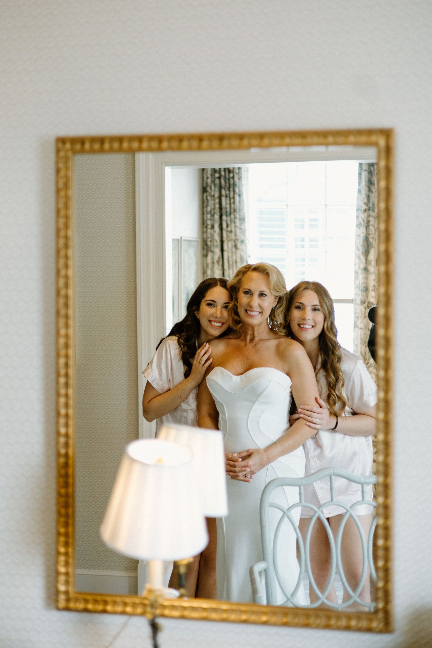 A wedding party gathers for a joyful mirror selfie in a brightly lit room with elegant decor and a gold-framed mirror.