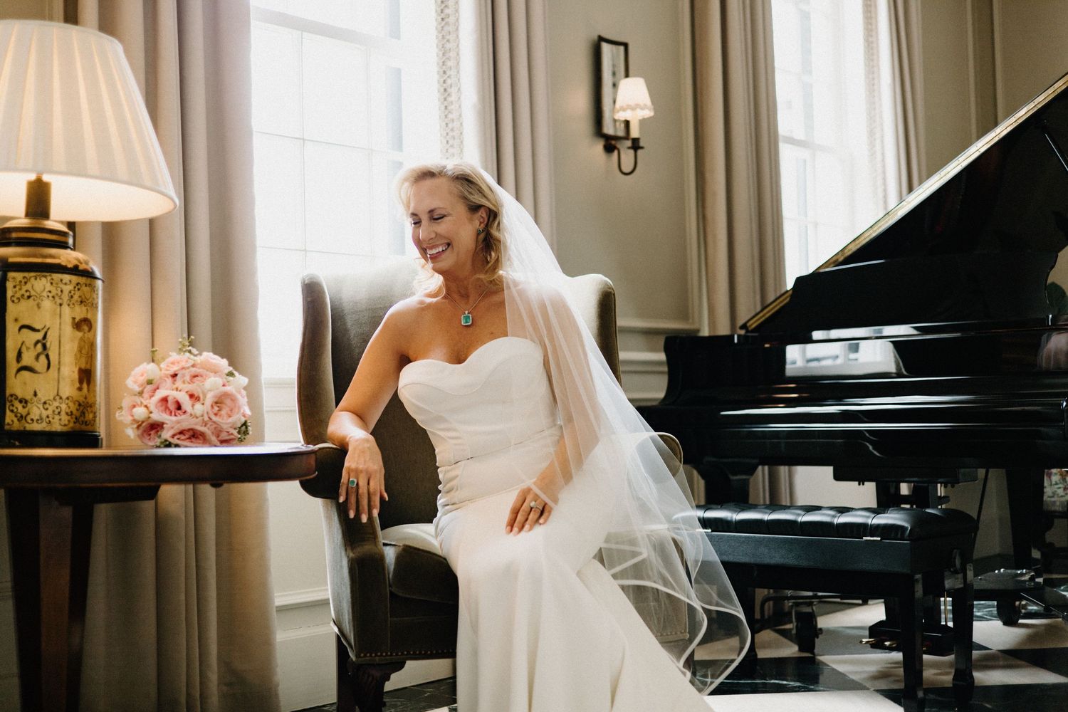 Elegant bride in a strapless white gown poses gracefully next to a grand piano in a luxurious room with cream decor.