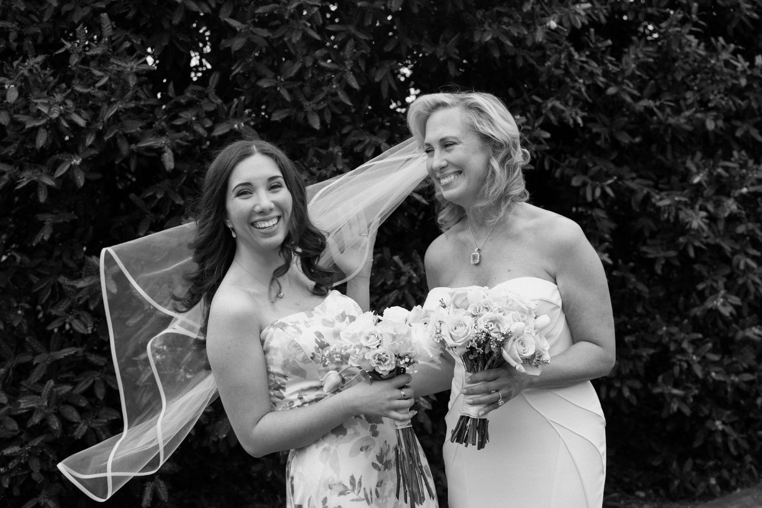 Black and white photo of two people in white dresses holding wedding bouquets outdoors.