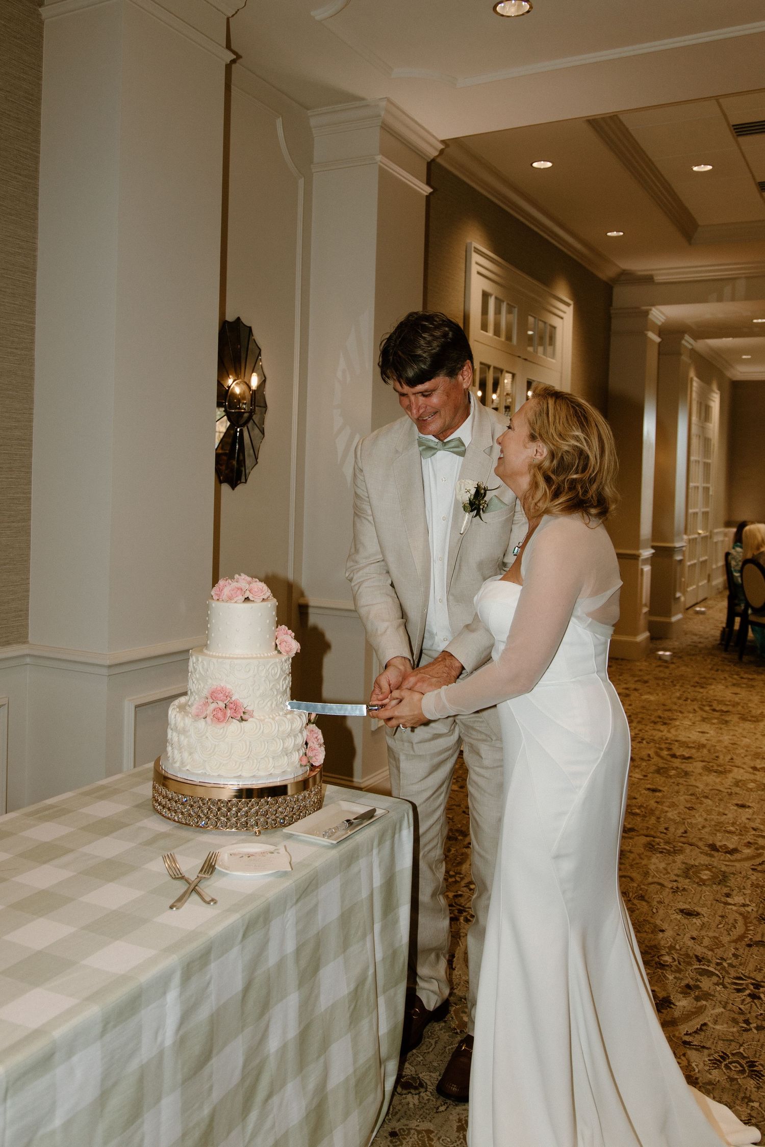 A couple in formal wedding attire cuts into their elegant three-tiered white wedding cake together at their reception.