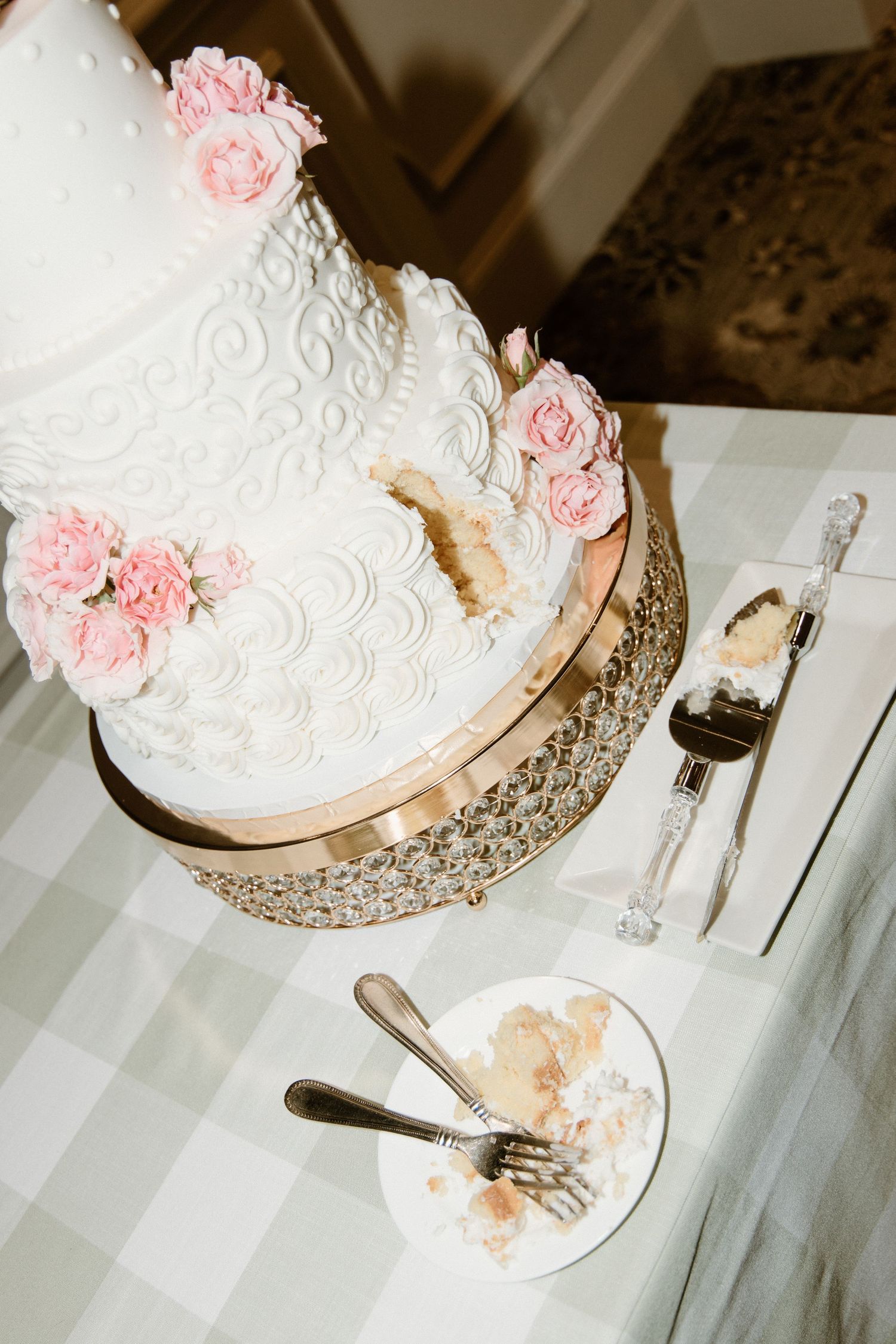 An elegant white wedding cake with pink sugar roses sits on a silver stand next to a partially eaten slice of cake.