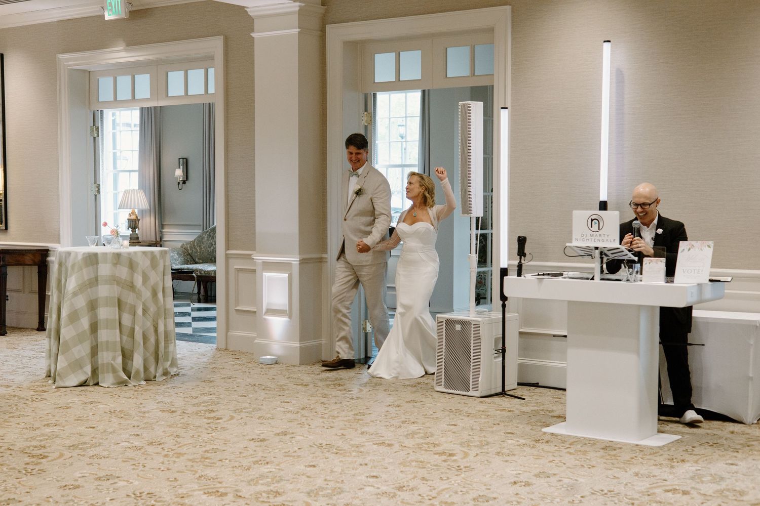 Two people in formal attire stand together in an elegant white ballroom with tall windows and reception desks.
