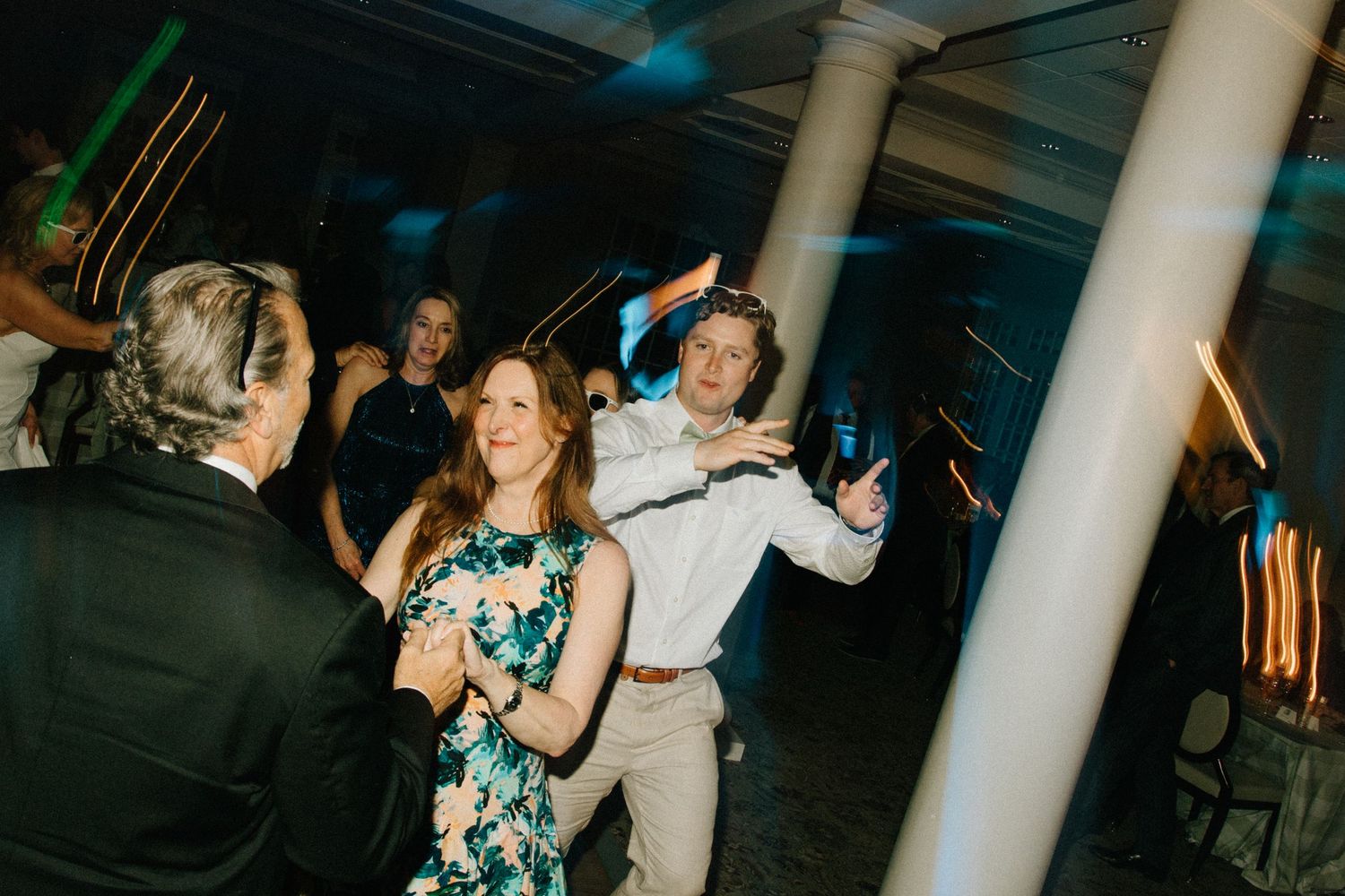 People dancing and having fun at a wedding reception in a venue with white columns and mood lighting.