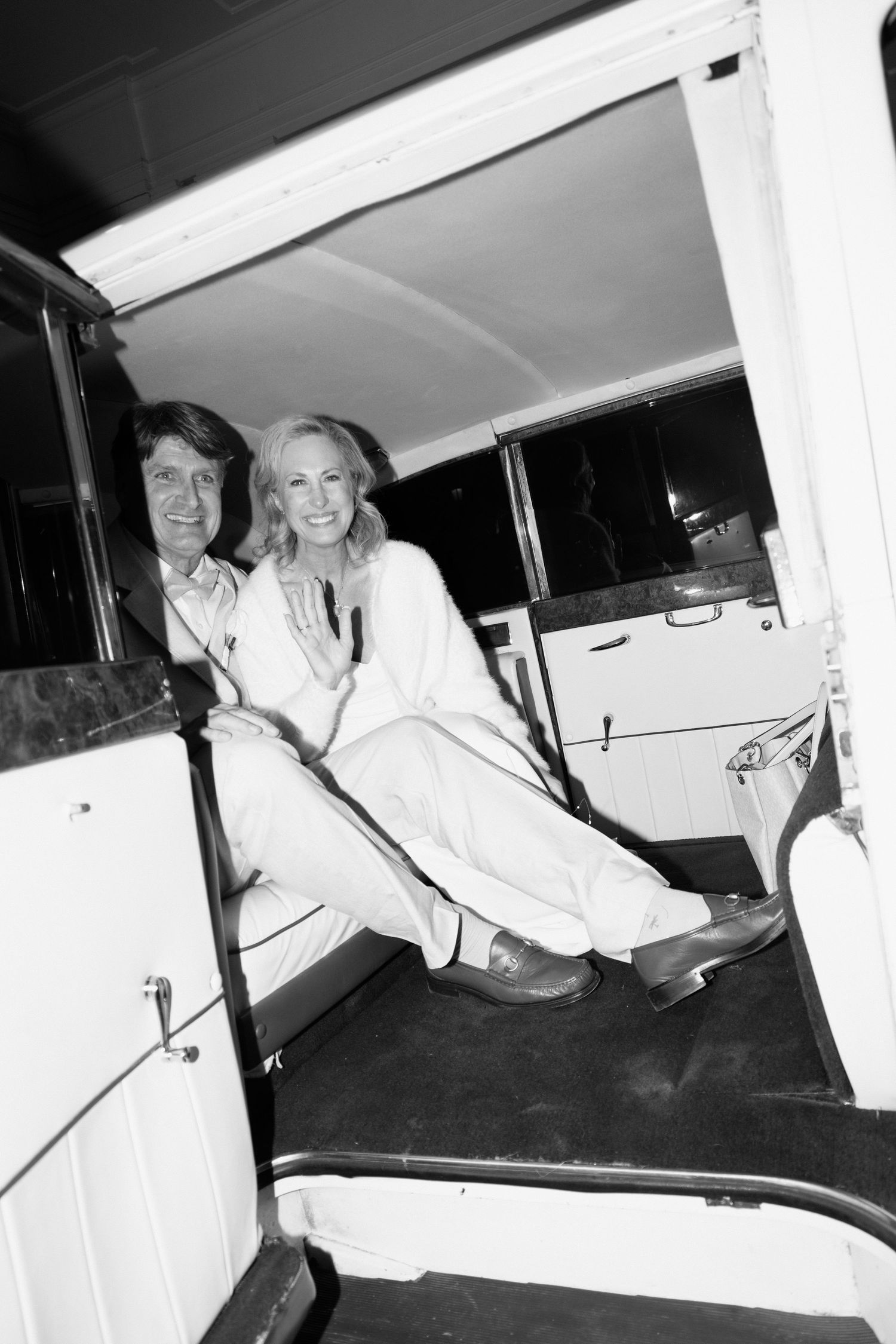 A black and white photograph showing two people sitting and laughing together in the back seat of a vintage car.
