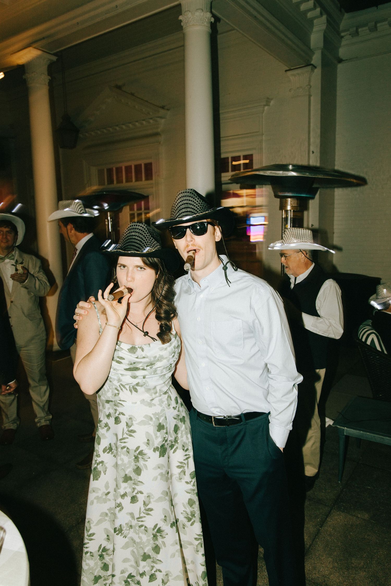 Party guests smoking cigars while wearing sunglasses and formal attire at an indoor event.