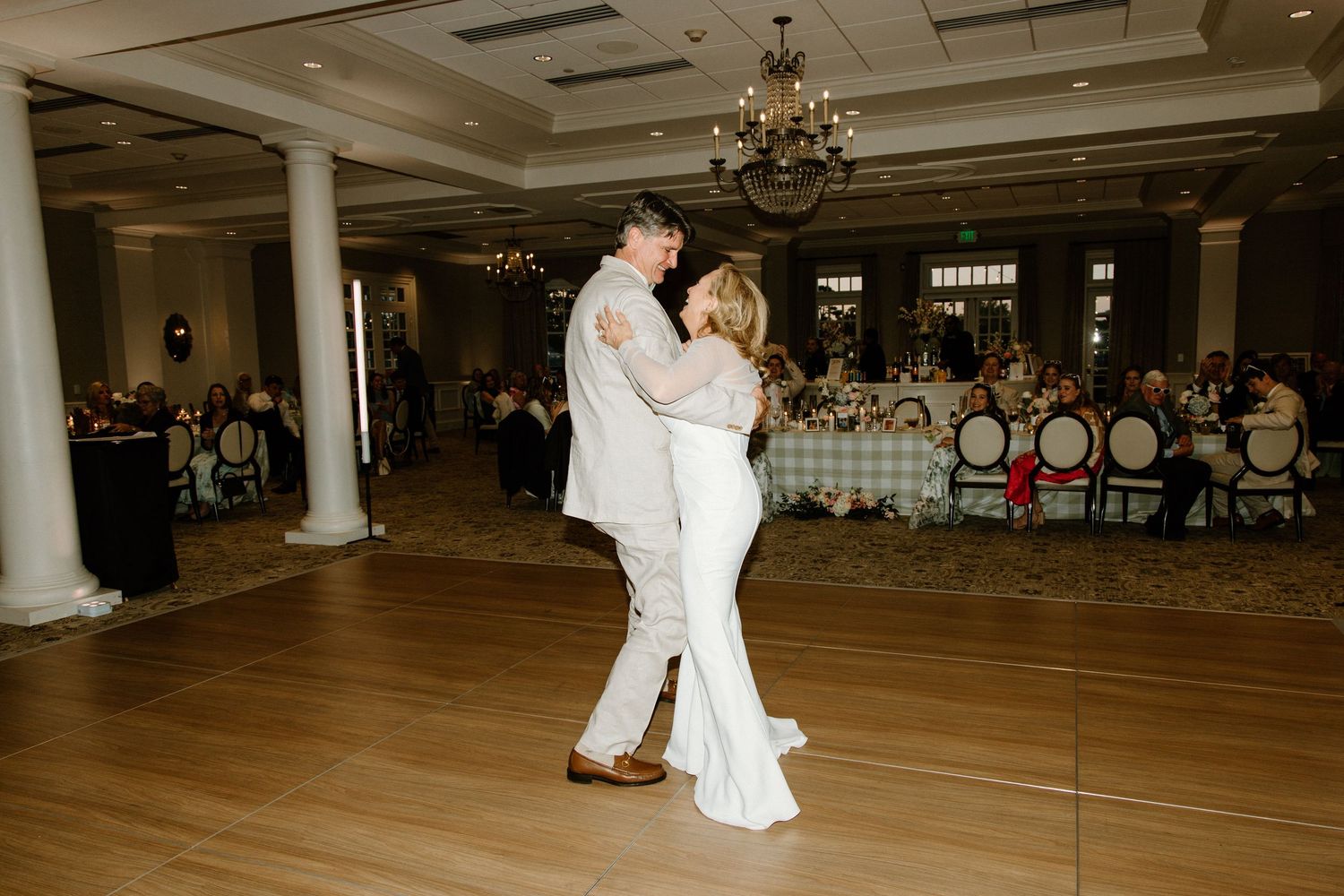 Two people in white outfits share an intimate first dance on a wooden floor at an elegant ballroom wedding reception.