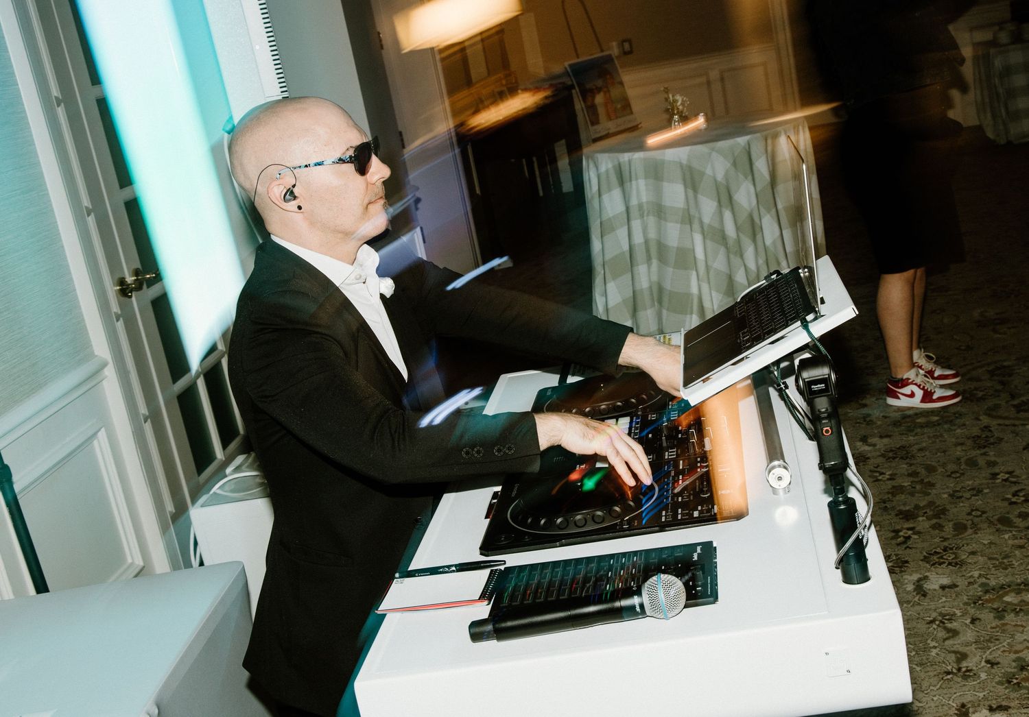 DJ in a tuxedo and sunglasses plays music at an event while operating electronic equipment at a counter setup.