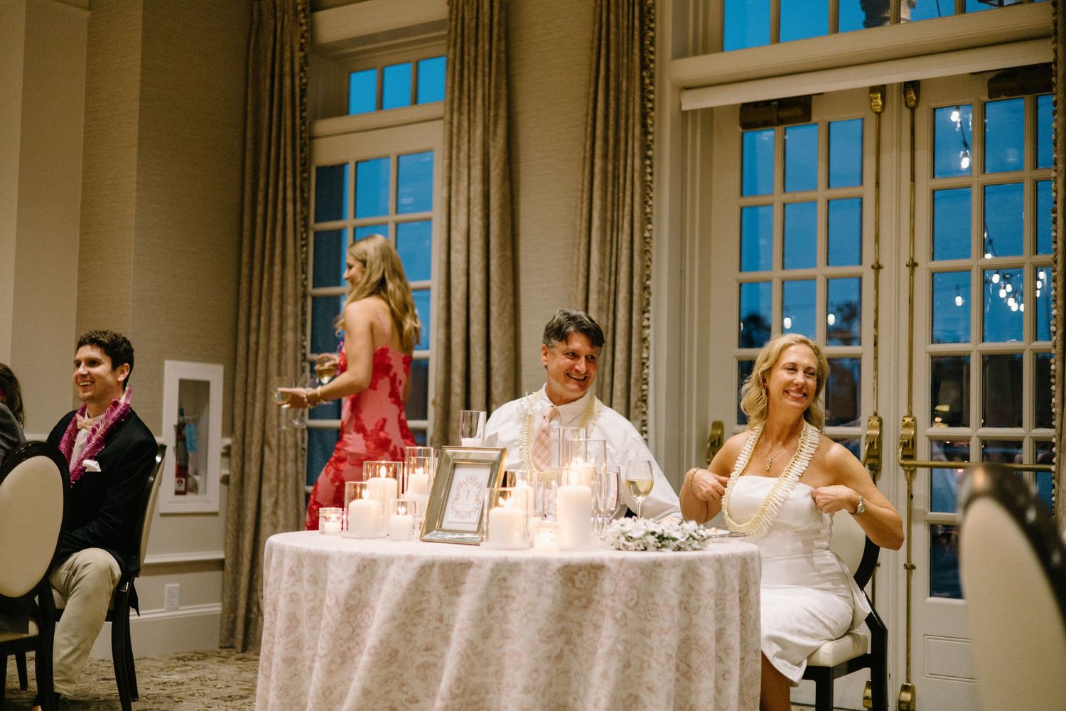 Wedding guests enjoying a casual moment at an elegantly decorated reception table in a grand ballroom with tall windows.