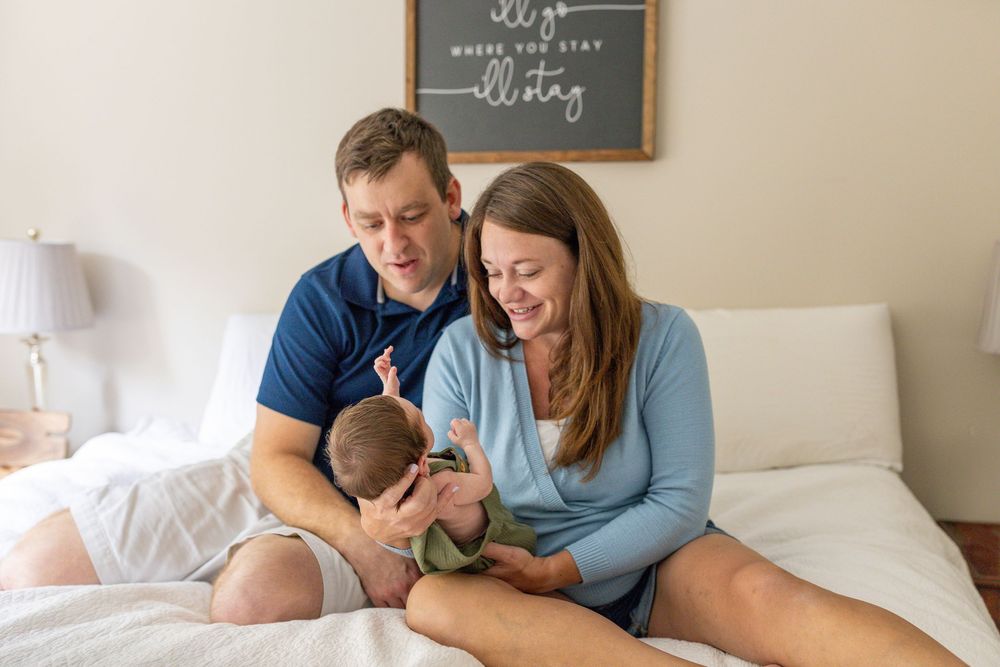 Parents and newborn baby sharing tender moments together on white bedding in a bright bedroom setting.