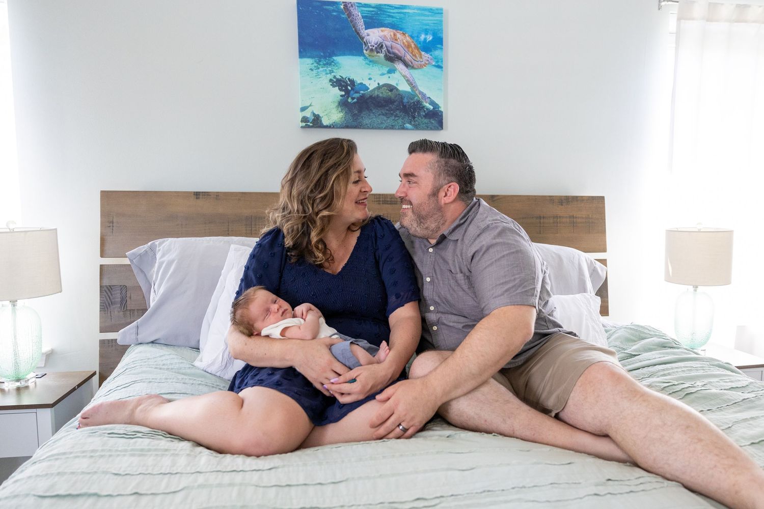 Couple relaxing together comfortably on white bedding in a bedroom with ocean artwork on the wall.