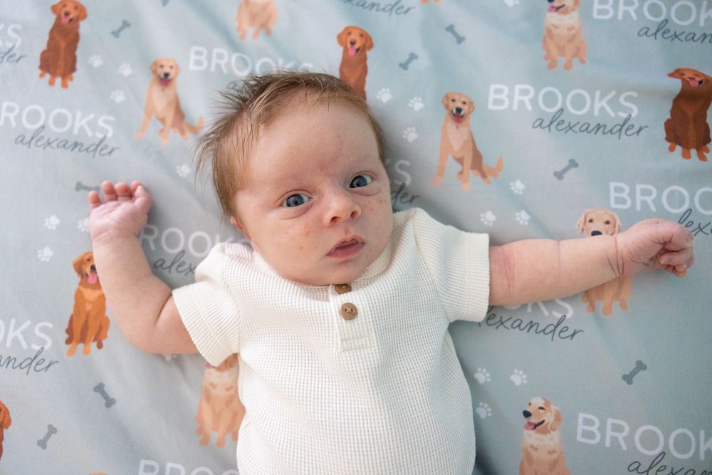 Baby lying on patterned bedding with teddy bear print in blue and orange tones.