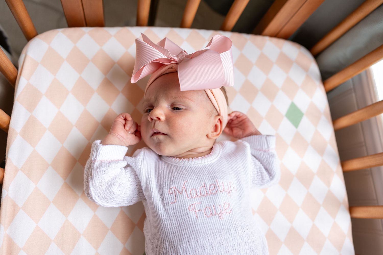 A sleeping baby wearing a white outfit and pink bow rests on a peach and white diamond-patterned crib sheet.