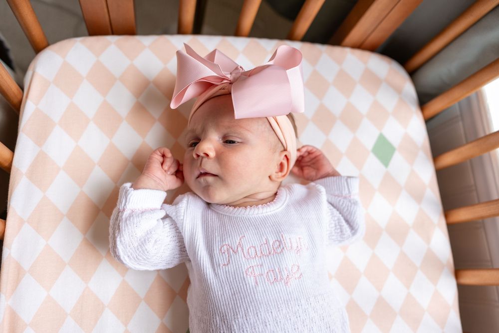 A sleeping baby wearing a white outfit and pink bow rests on a peach and white diamond-patterned crib sheet.