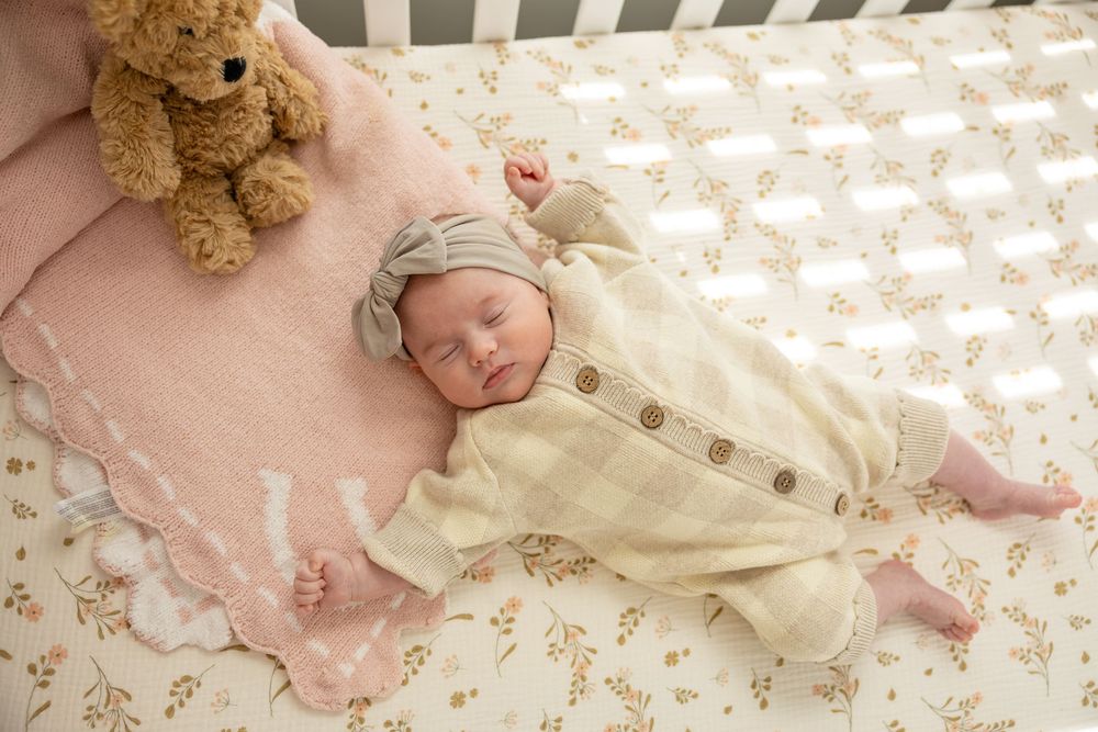 Newborn baby sleeping peacefully in a crib wearing a plaid outfit and hat, beside a pink blanket and teddy bear.