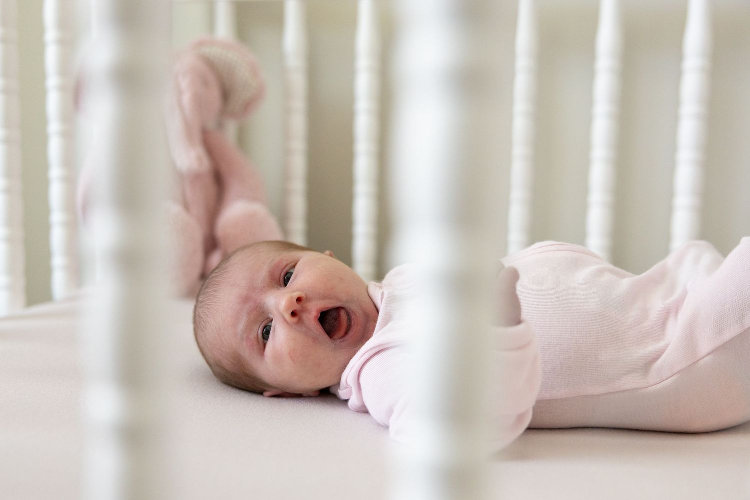 Baby yawning while lying in white crib with slats visible in foreground.
