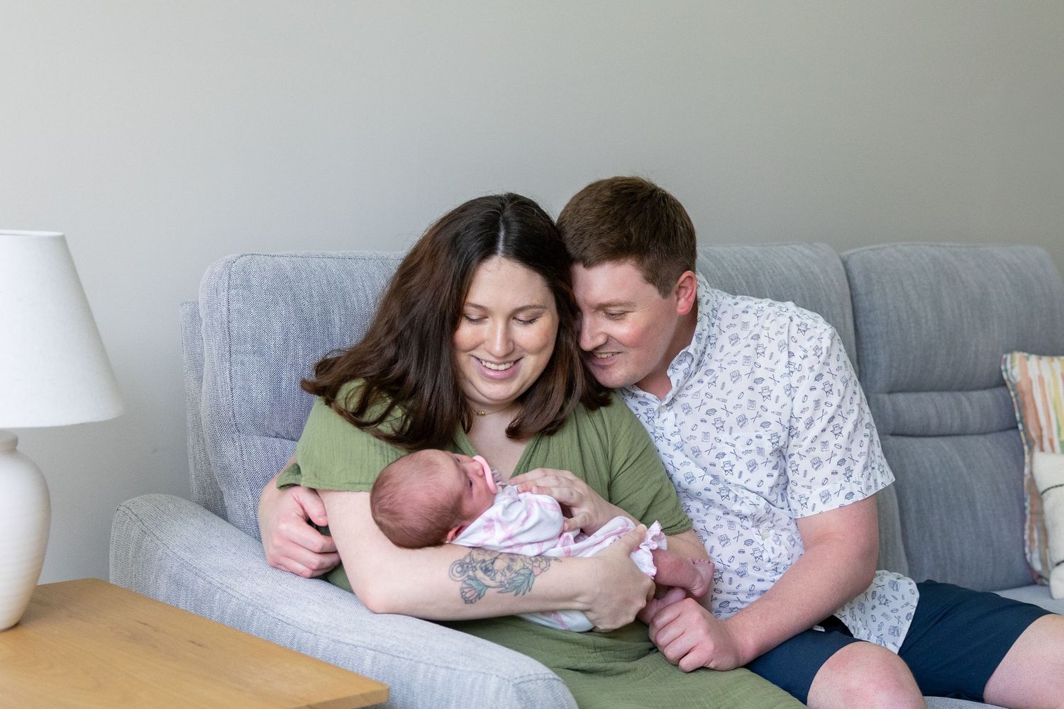 A couple sits together on a gray couch while tenderly cradling their newborn baby.