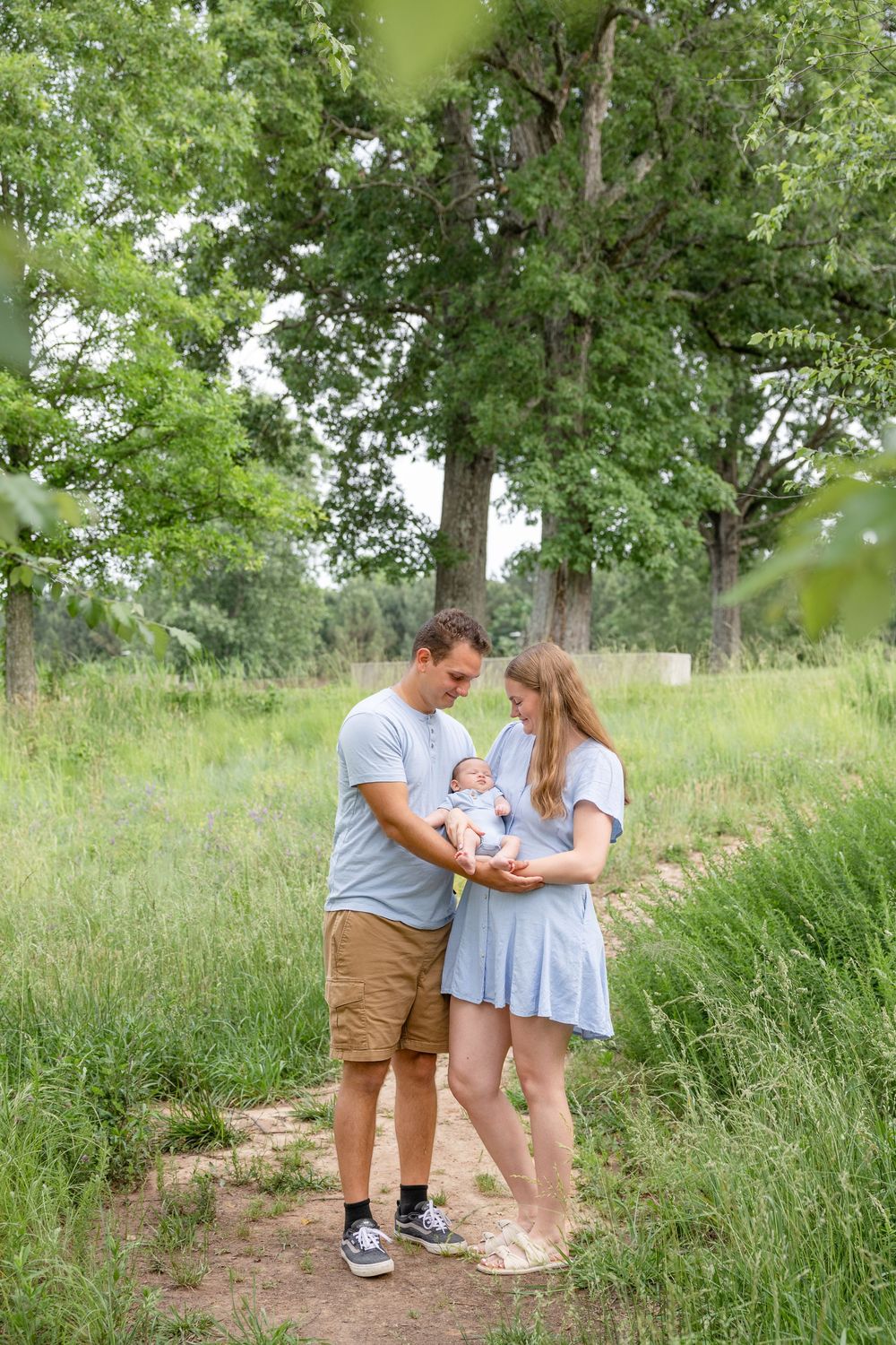 A couple stands together on a dirt path surrounded by tall grass and trees while holding a baby on a summer day.