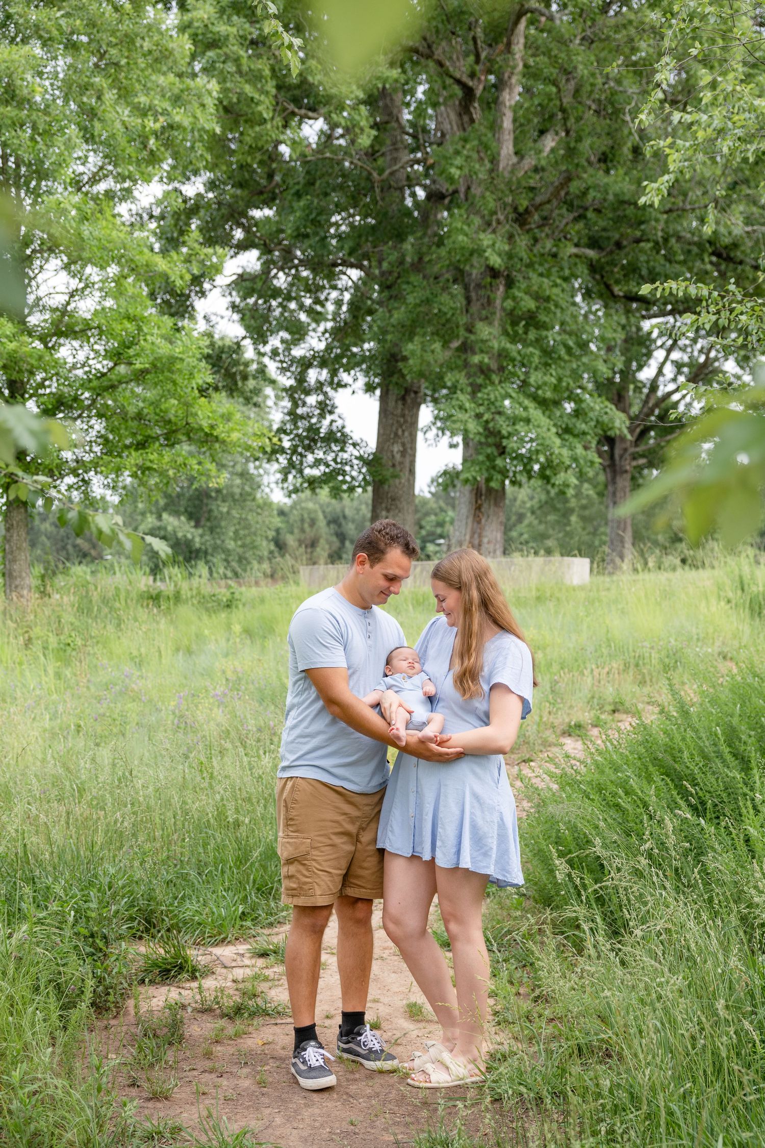 A couple stands together on a dirt path surrounded by tall grass and trees while holding a baby on a summer day.