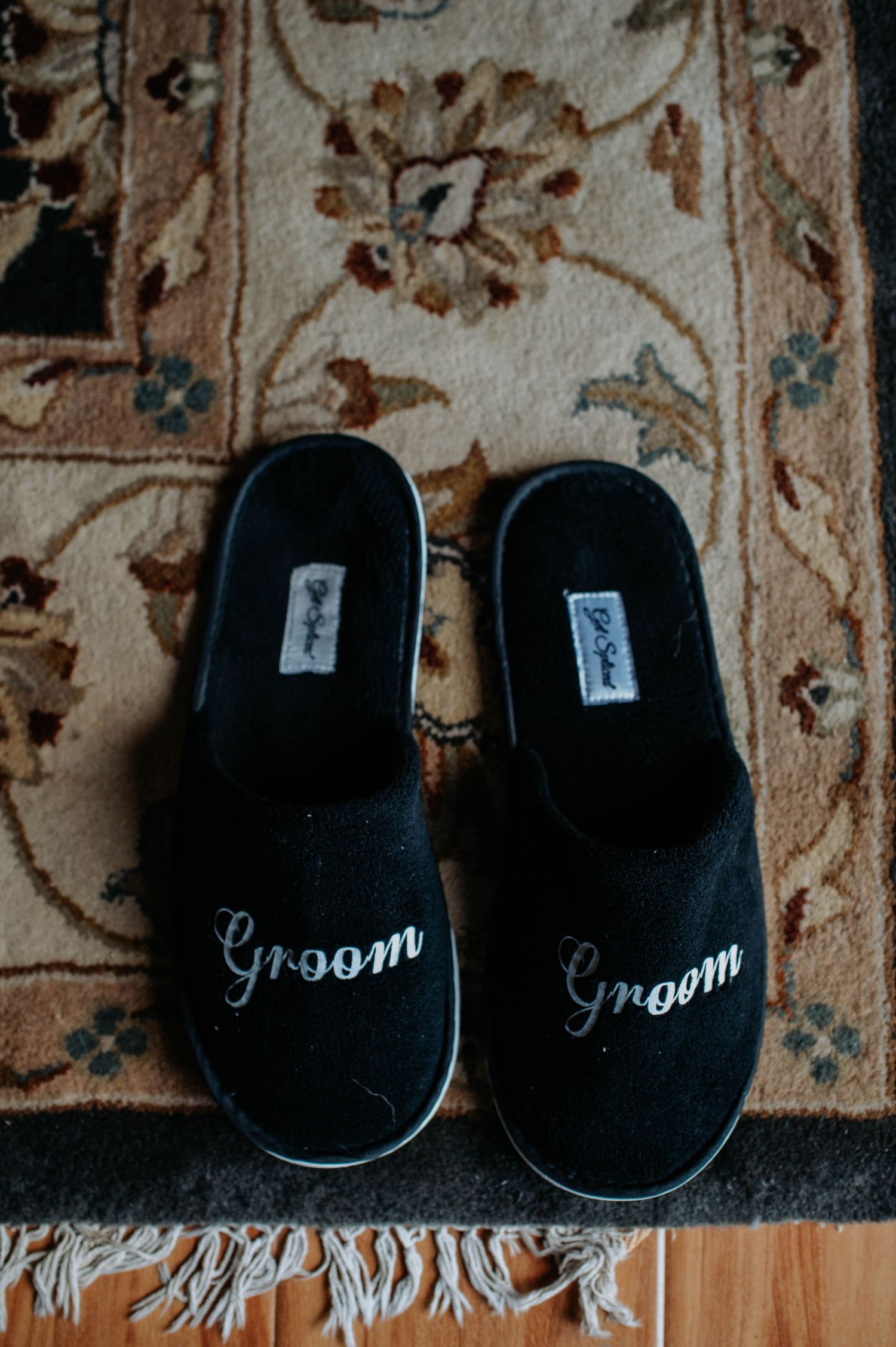 A pair of black velvet slippers with 'groom' embroidered text arranged on a vintage patterned carpet.