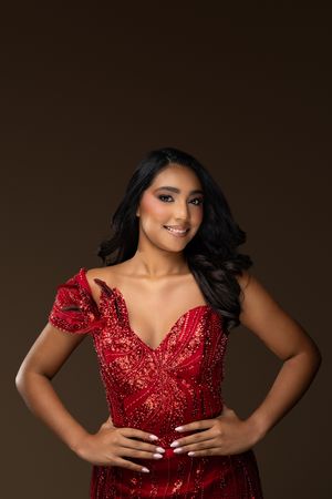 Young woman in red sequin gown with white nails poses confidently against dark brown background.