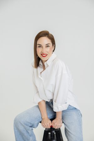 A person in a white button-down shirt and jeans sits casually on a light gray background.