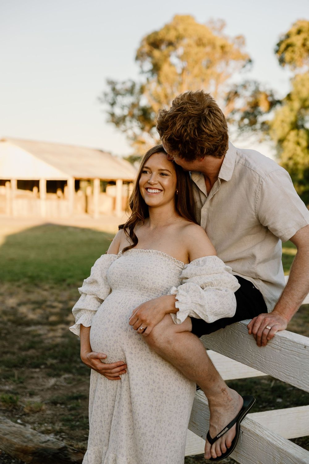 Romantic maternity photoshoot with couple in white dress and casual attire by wooden fence at sunset.