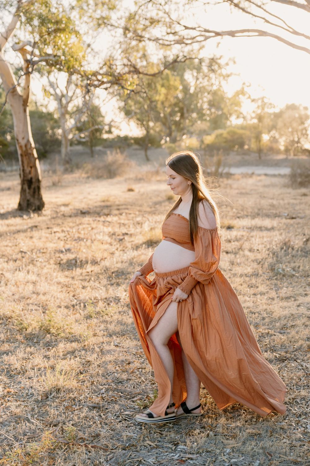 A sunset maternity photoshoot outdoors with a flowing rust-colored dress against a backdrop of eucalyptus trees.