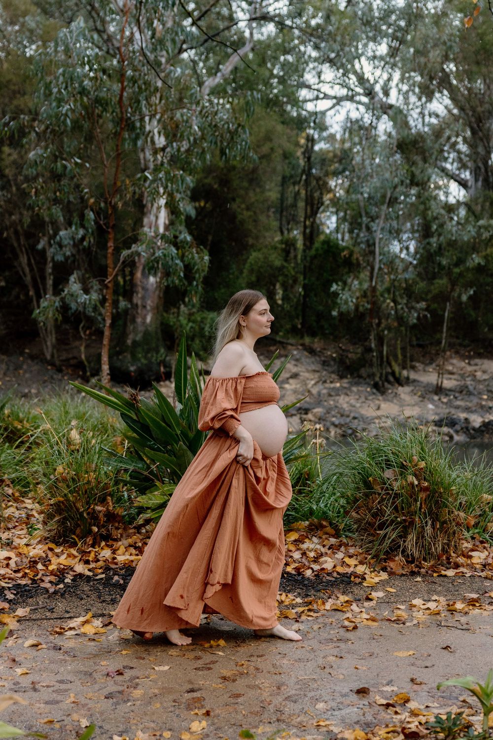 A person in a flowing terracotta-colored dress walks along an autumn path surrounded by eucalyptus trees and fallen leaves.
