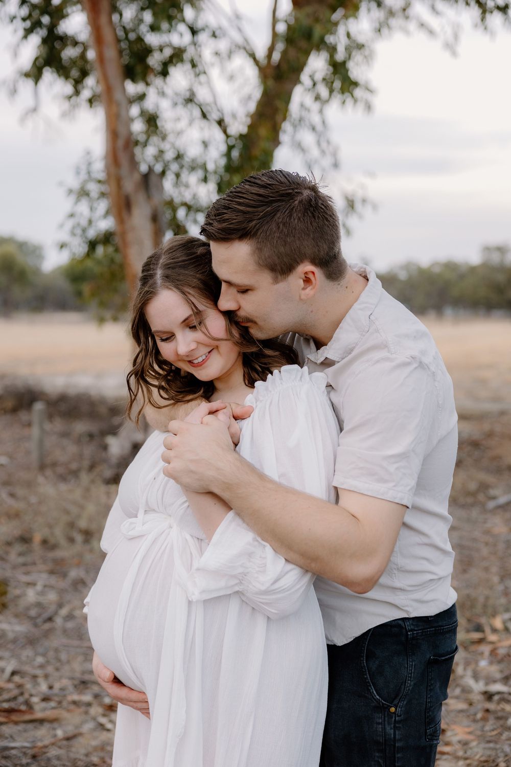 A couple shares an intimate embrace outdoors in casual white and dark clothing against a natural backdrop.