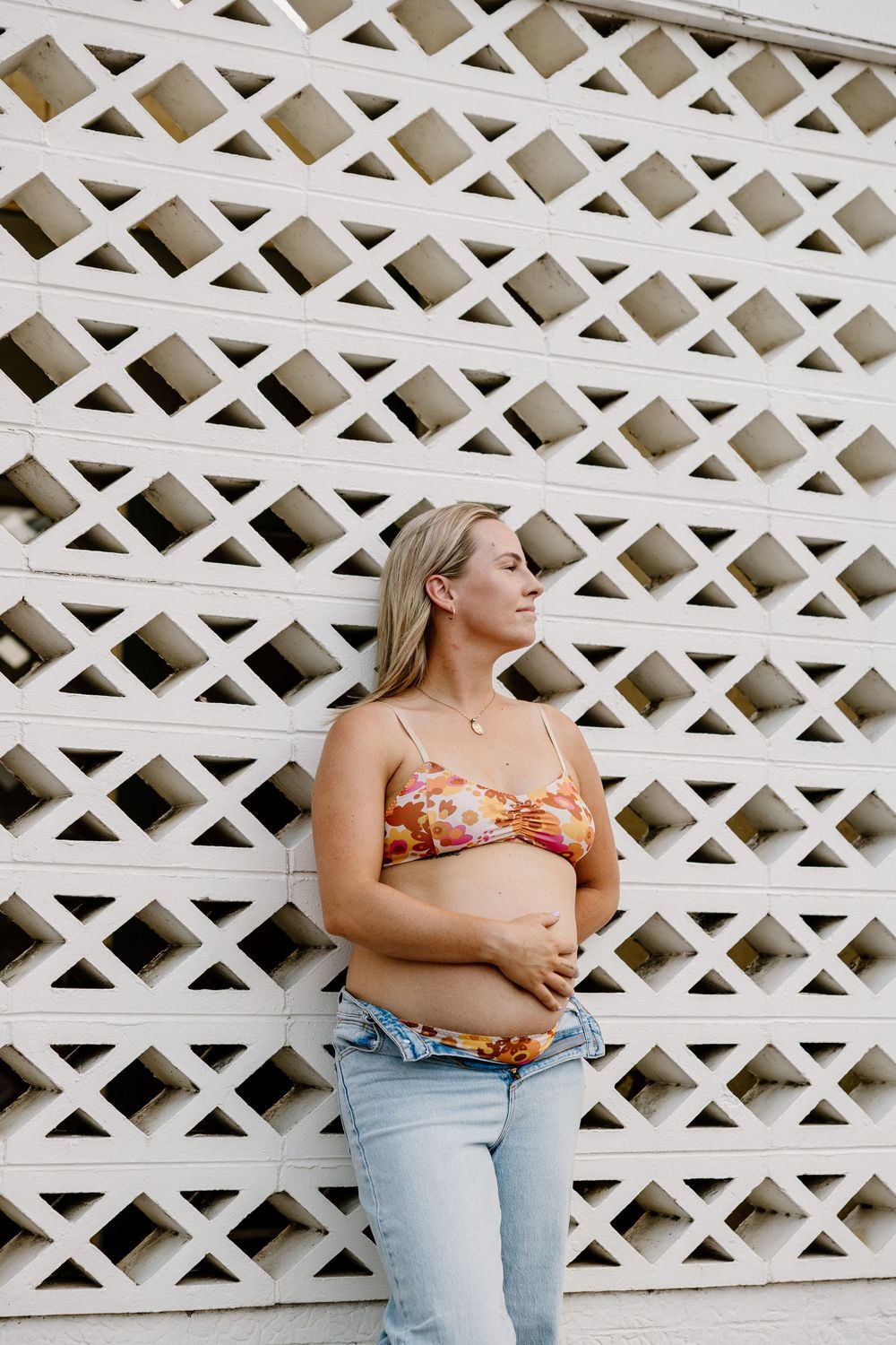 A person wearing a floral top and jeans poses against a white lattice wall with geometric patterns.
