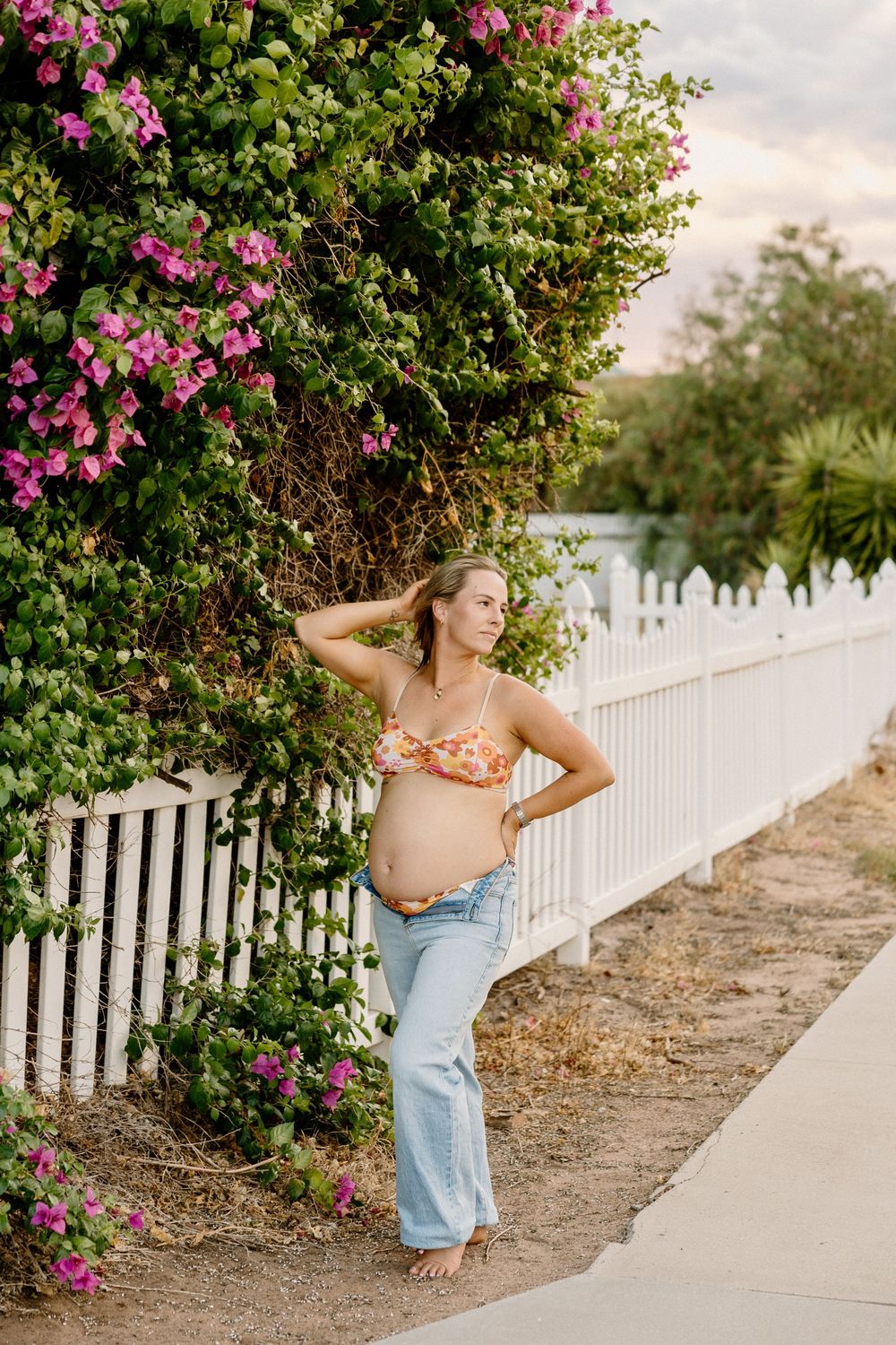 A pregnant woman in jeans and floral top poses beside a white picket fence and pink flowering bush.