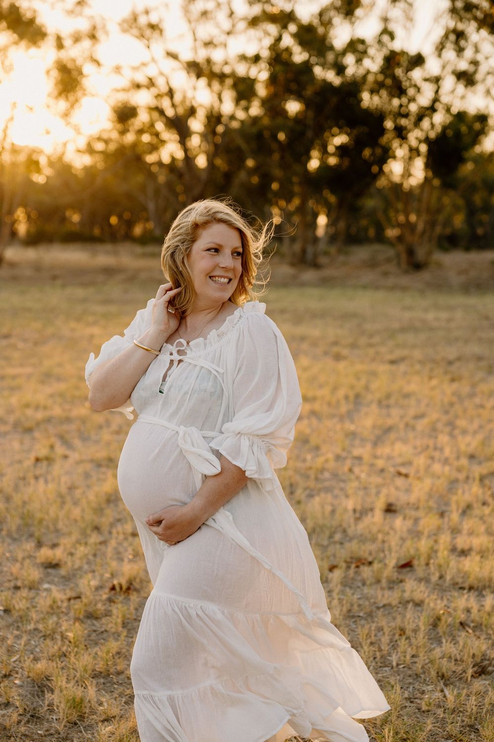 Pregnant woman in flowing white dress stands in golden sunlit field at sunset while her hair blows in the breeze.