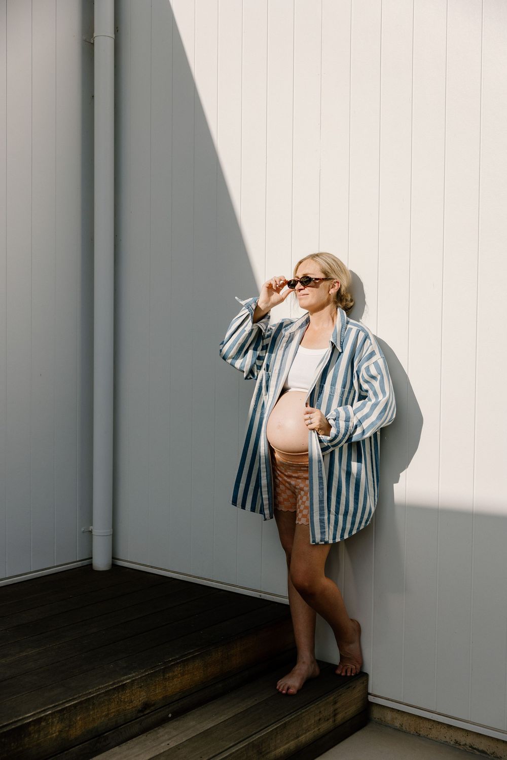 Expectant person in striped oversized shirt and shorts poses against white wall with dramatic lighting and shadows.