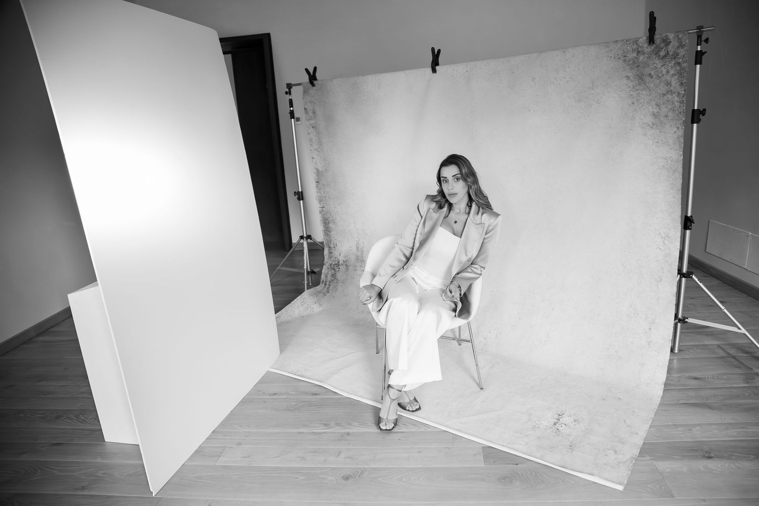 Behind the scenes black and white photo of a model seated in white dress against a studio backdrop during photoshoot setup.