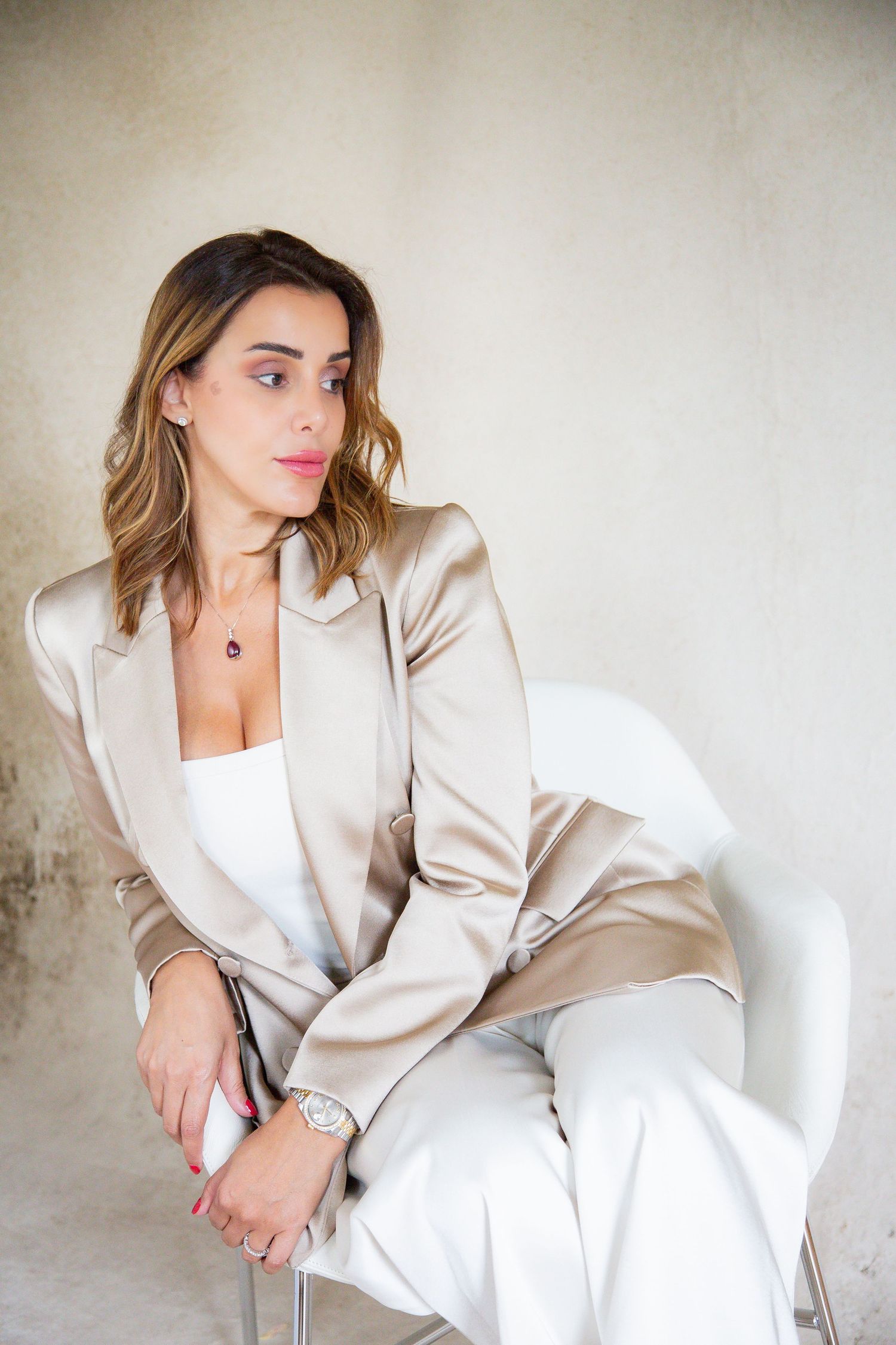 Woman in cream satin blazer and white outfit poses elegantly on white chair.