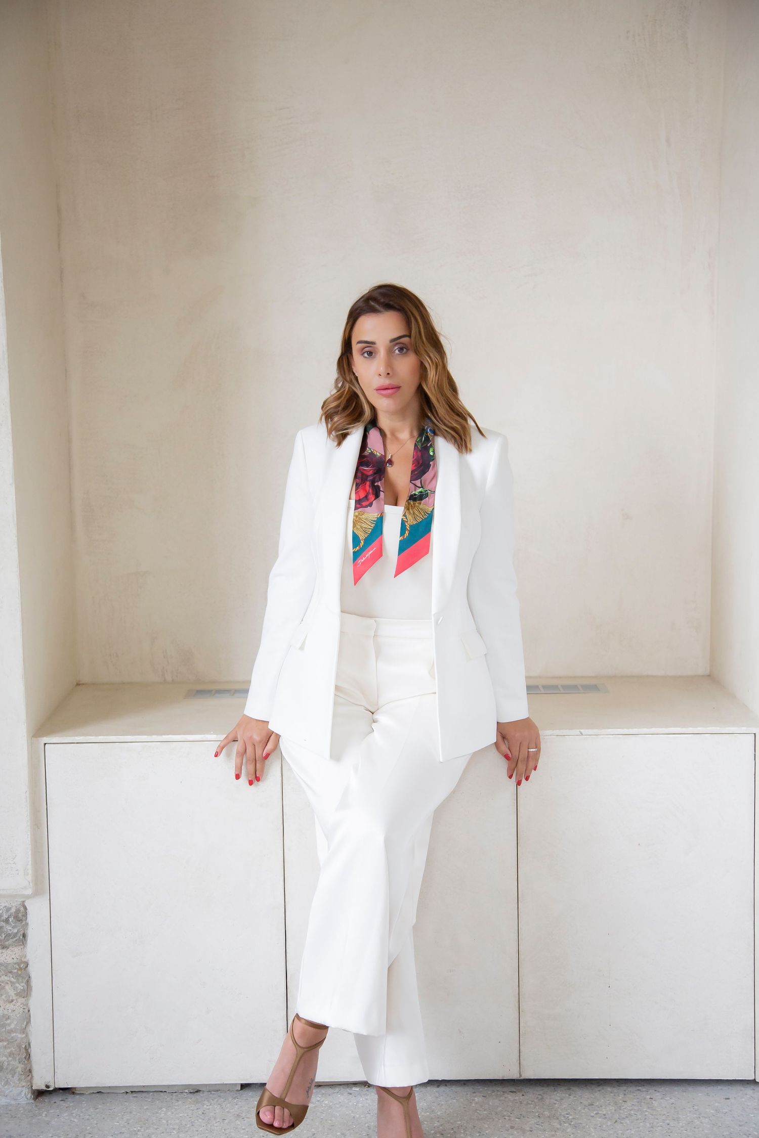 Woman in white pantsuit and vibrant patterned scarf poses against minimalist backdrop.