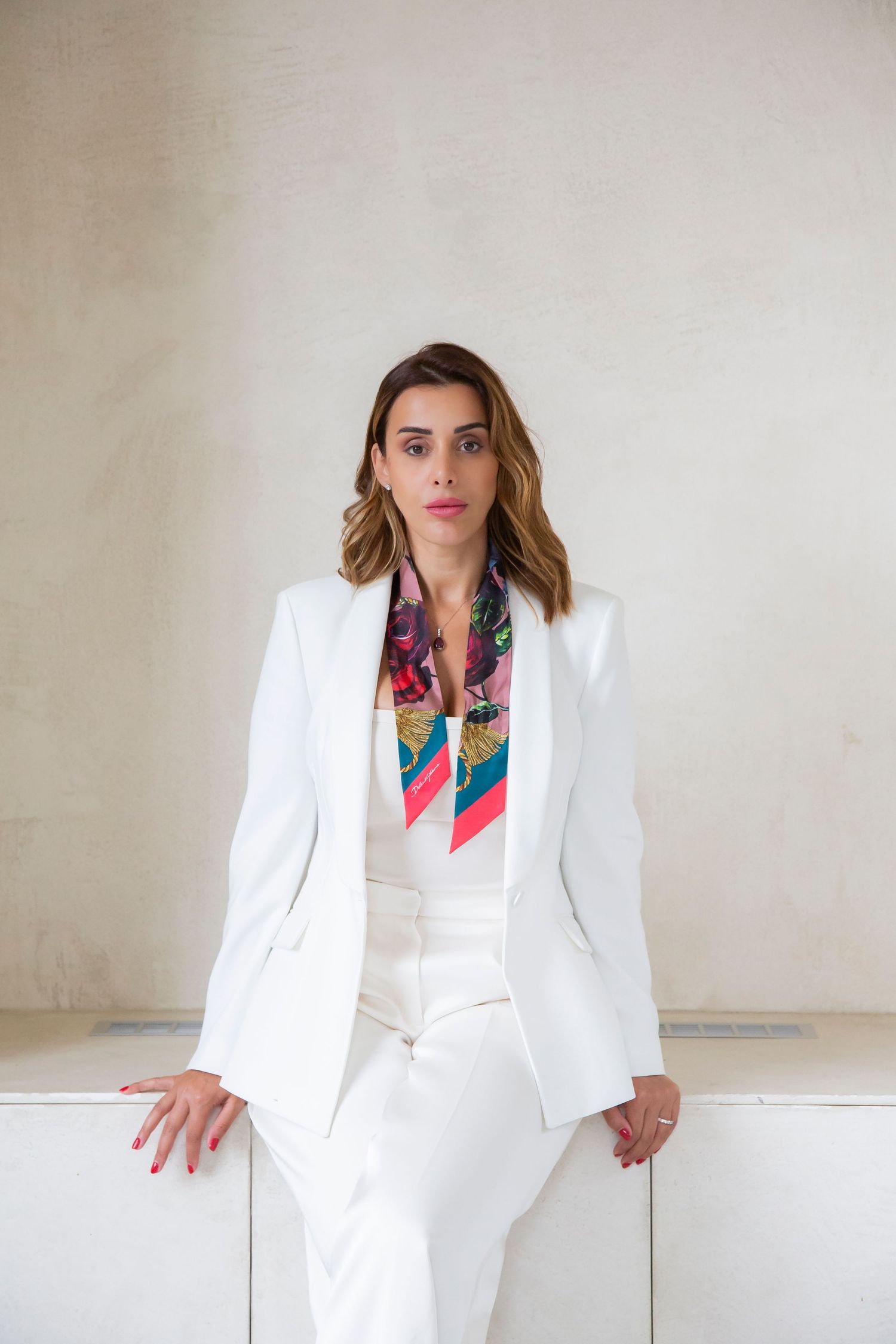 Model in crisp white suit and vibrant silk scarf poses against neutral wall in professional photoshoot.