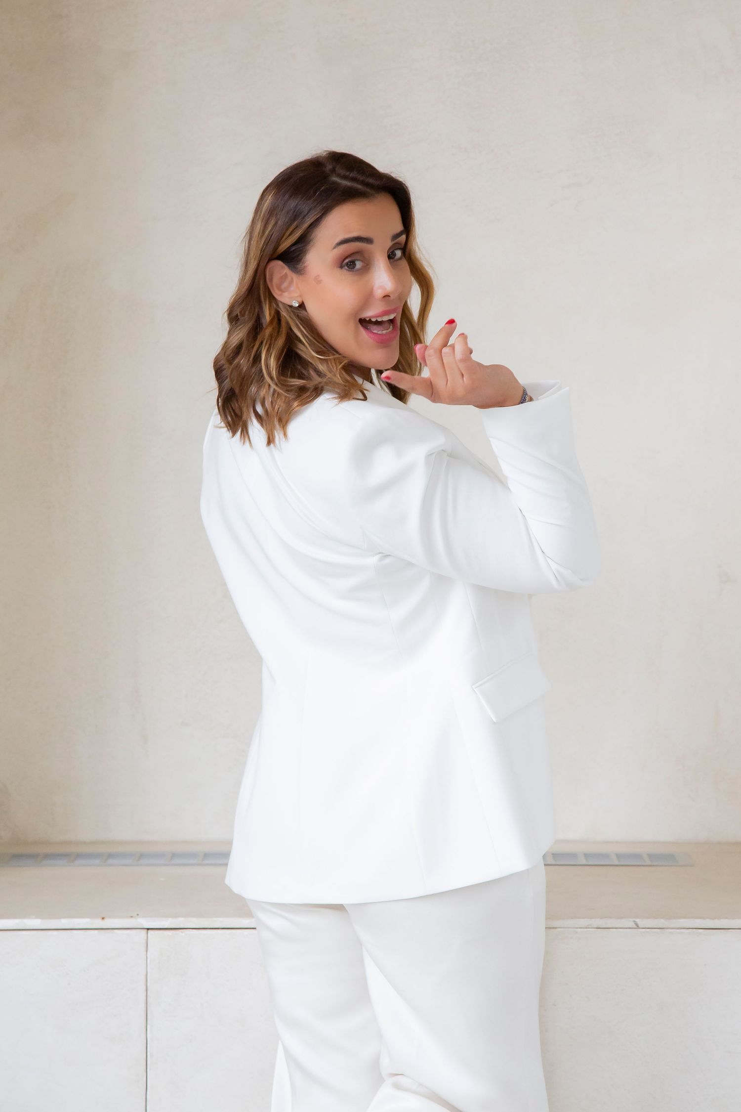 Person wearing all white business attire poses playfully against a light colored wall.