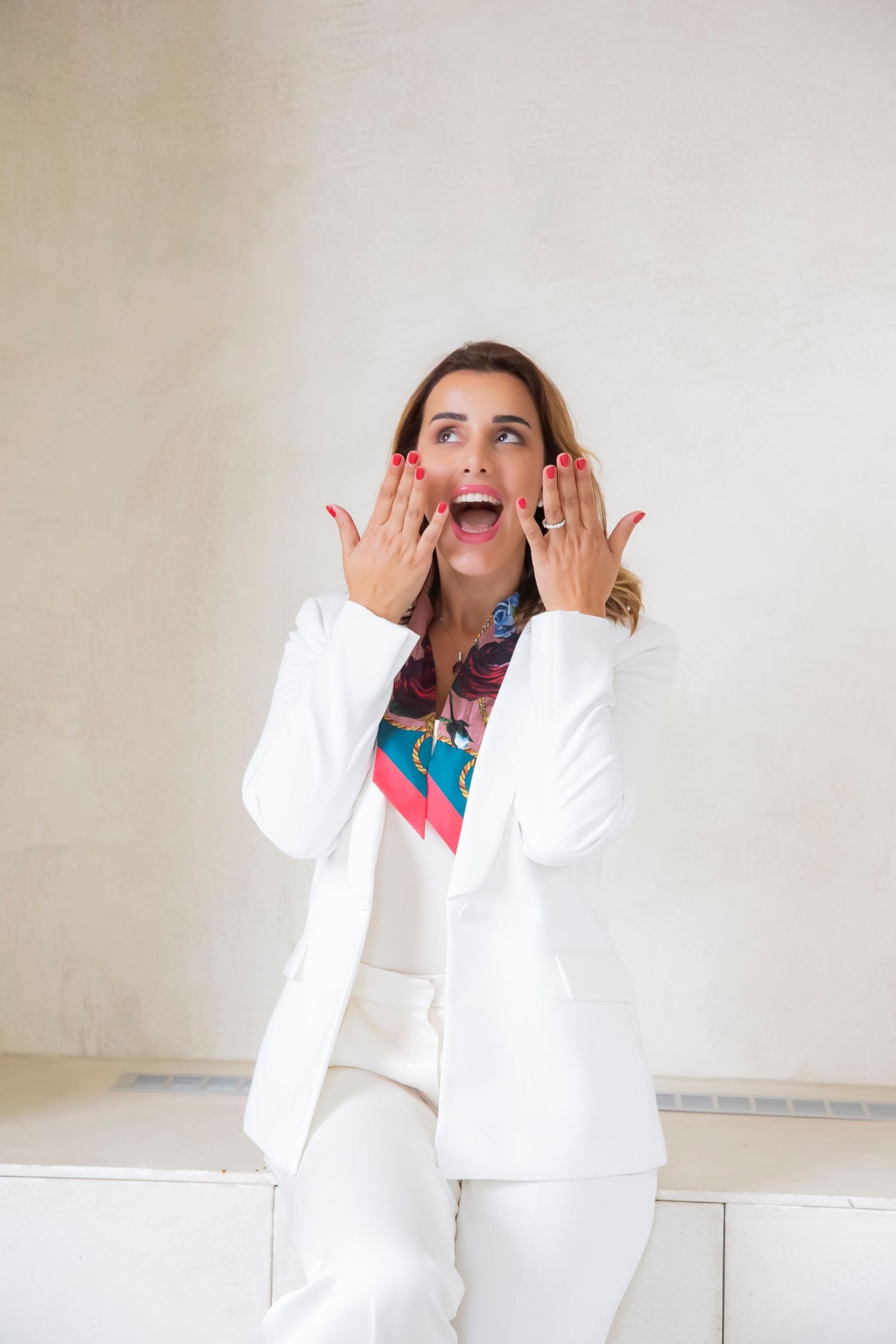 Person in crisp white suit and colorful top shows excited expression with hands raised near face against light wall.