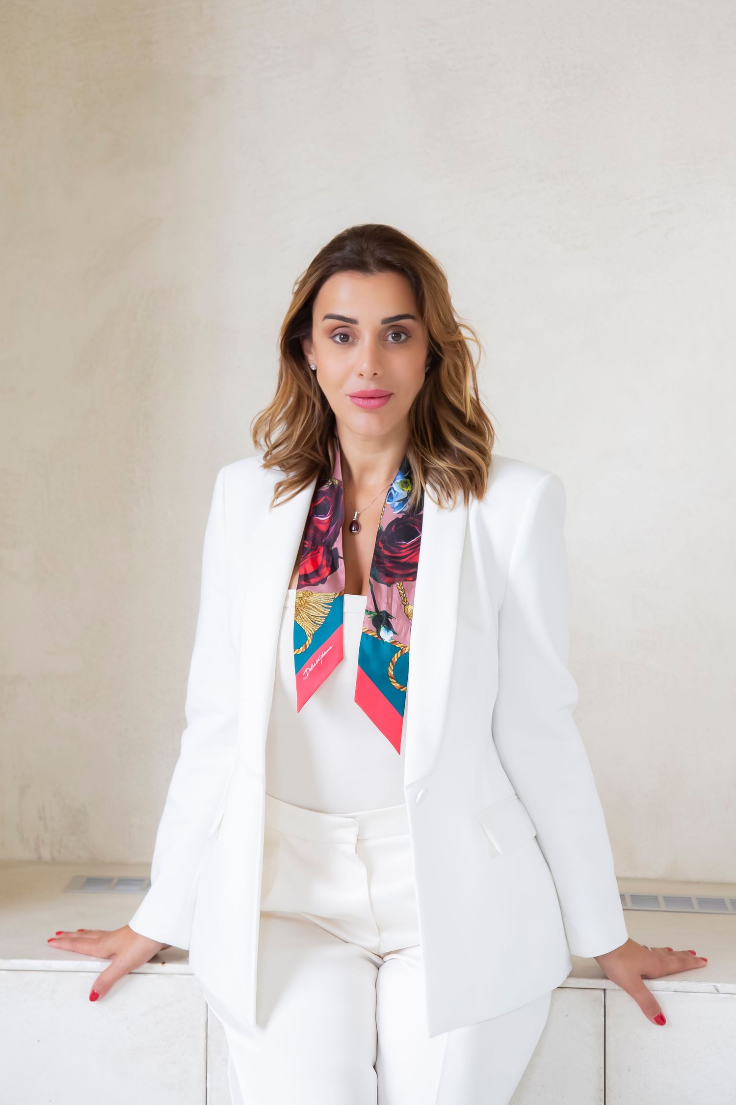 Professional portrait of a person in an all-white pantsuit ensemble with a colorful silk scarf leaning against a white surface.