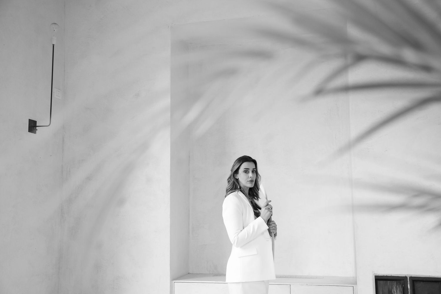 Black and white photo shows a person in white clothing standing against a wall with palm tree shadows cast on the surface.