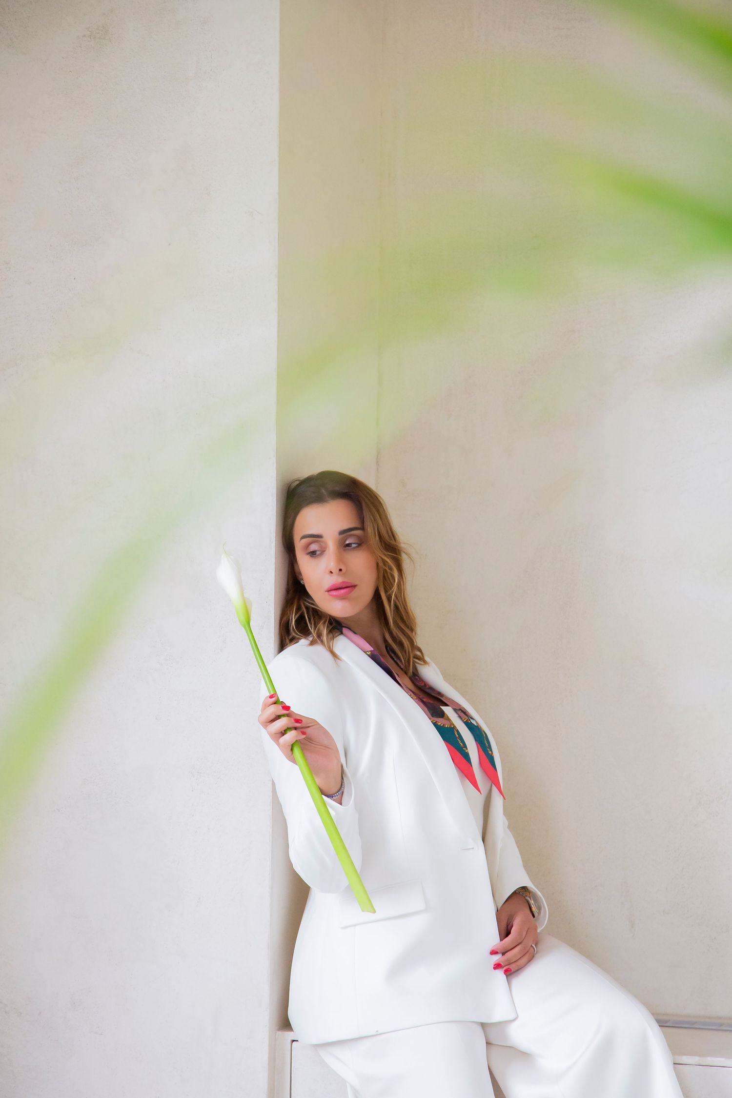 Figure in an all-white suit stands elegantly against a light wall holding a single calla lily flower.