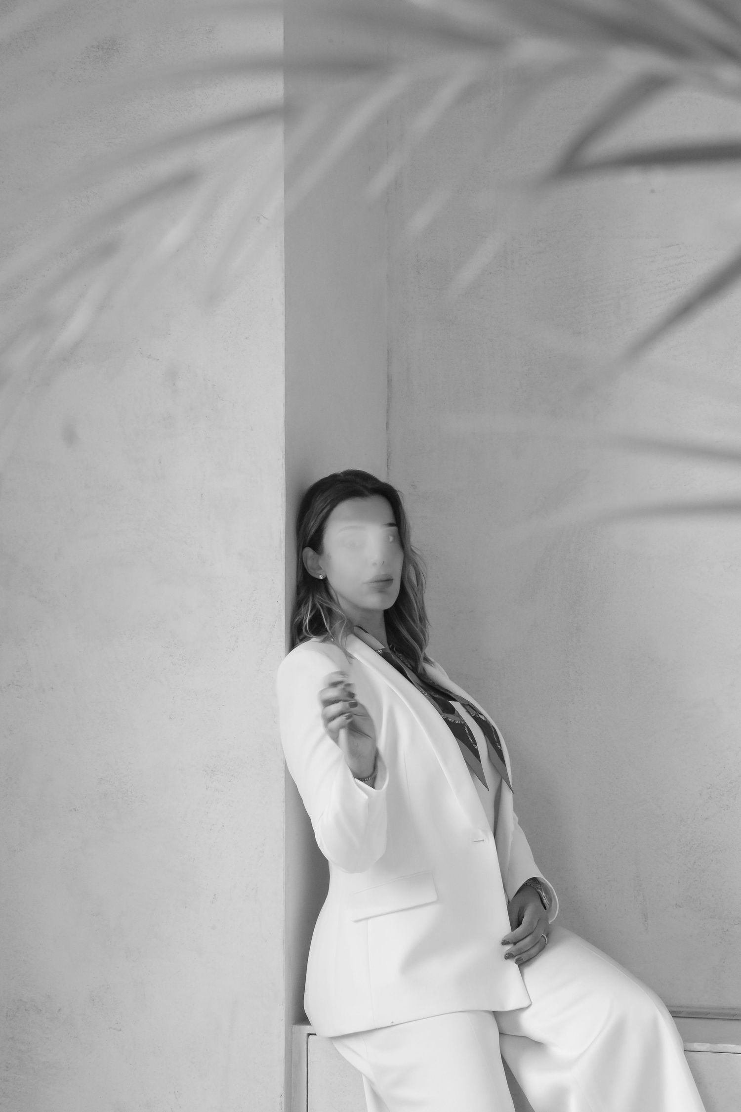 A dramatic black and white photo of someone in a white suit posing against a wall with palm frond shadows.