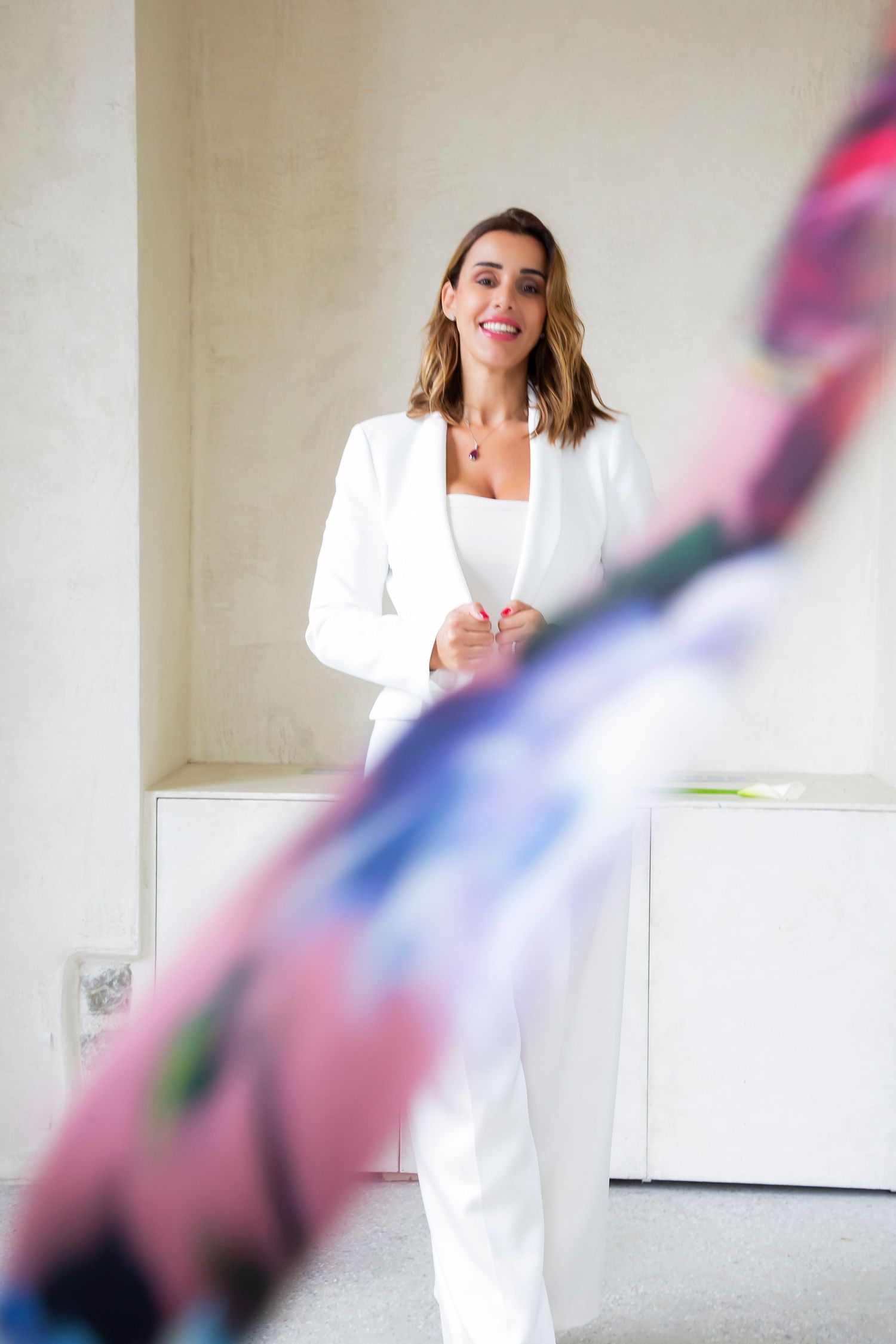 Professional fashion model wearing crisp white pantsuit poses confidently against light wall with blurred foreground detail.