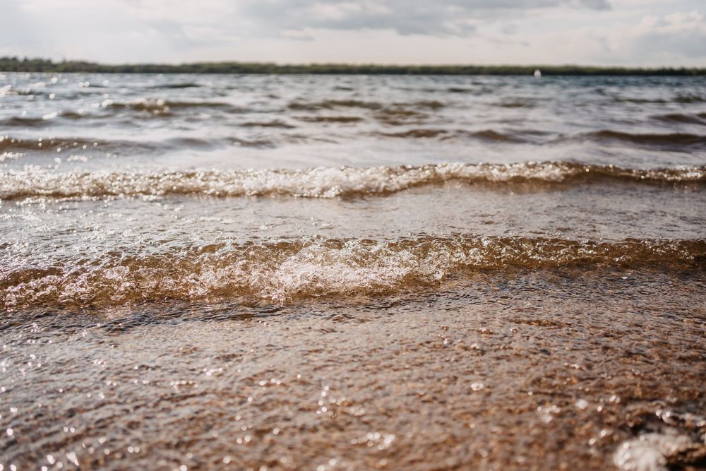 Brown foamy waves roll onto a sandy beach shore with choppy waters stretching to the cloudy horizon.